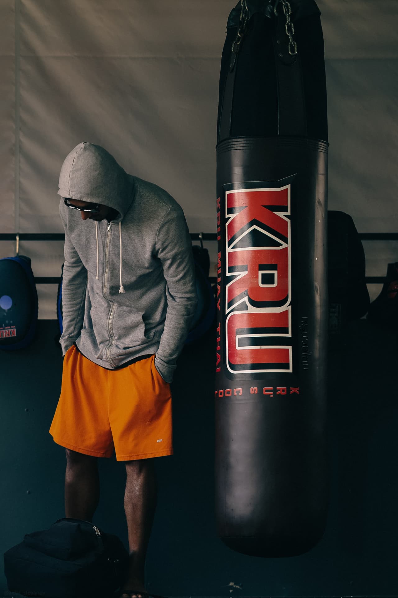 A man in a grey hoodie resting against a KRU heavy bag