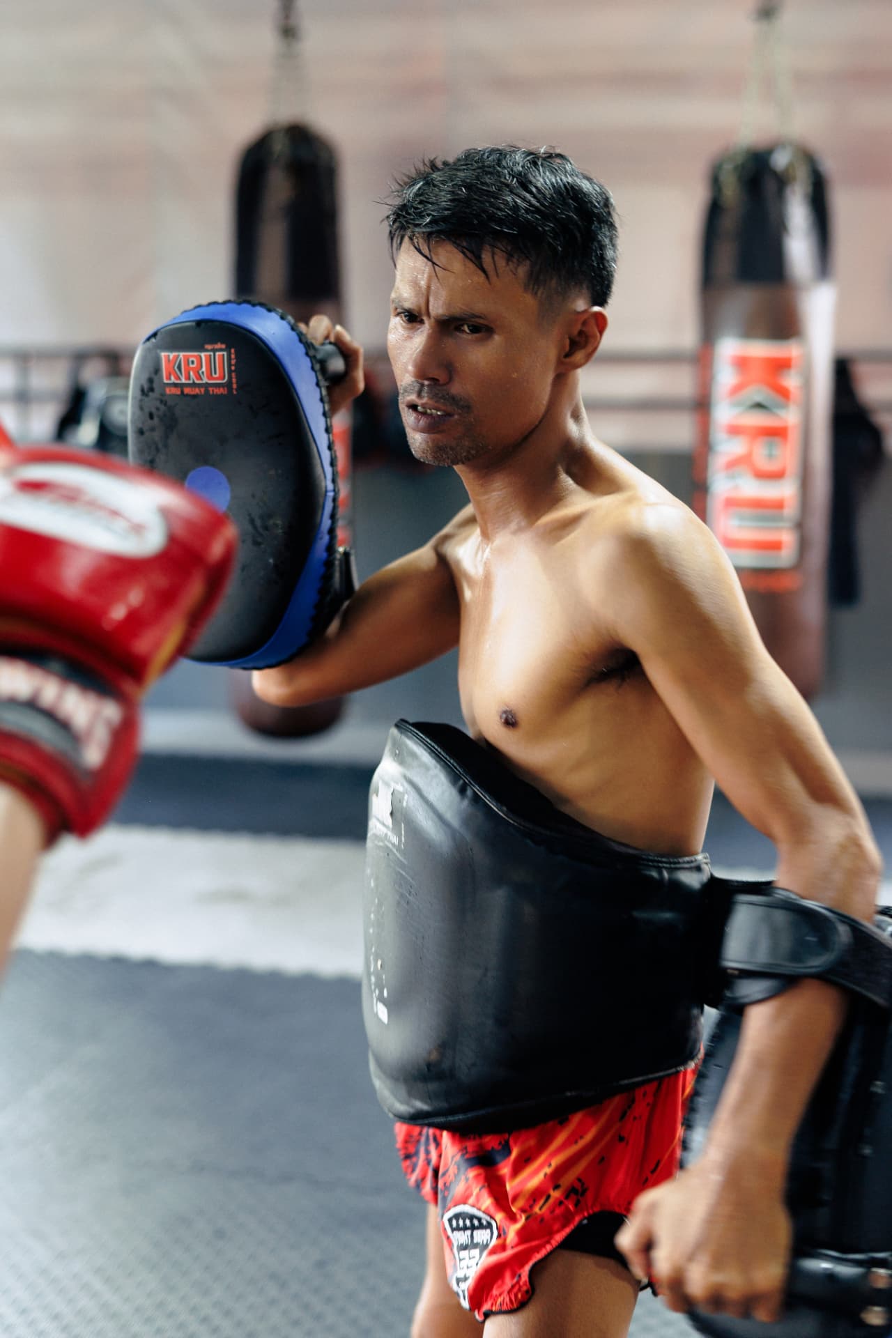 Kru Yai holding a blue focus pad and a body shield on the gym floor